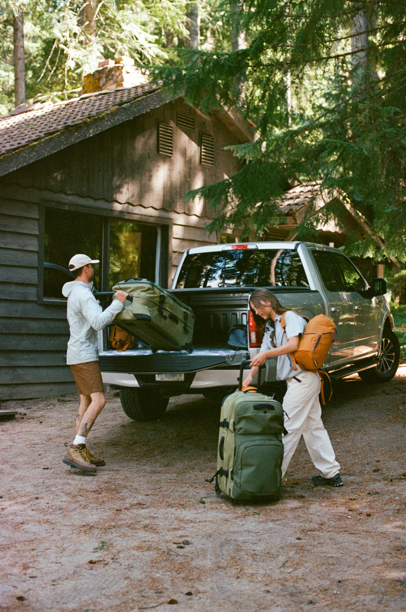 Two people loading and unloading large green hiking backpacks from the back of a pickup truck in a forest campsite setting with a wooden cabin.