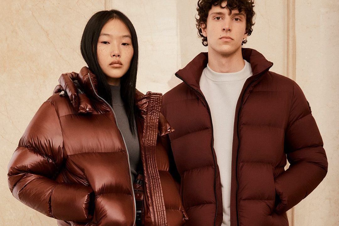 Two people wearing brown puffer jackets against a beige wall. Photo by Moose Knuckles