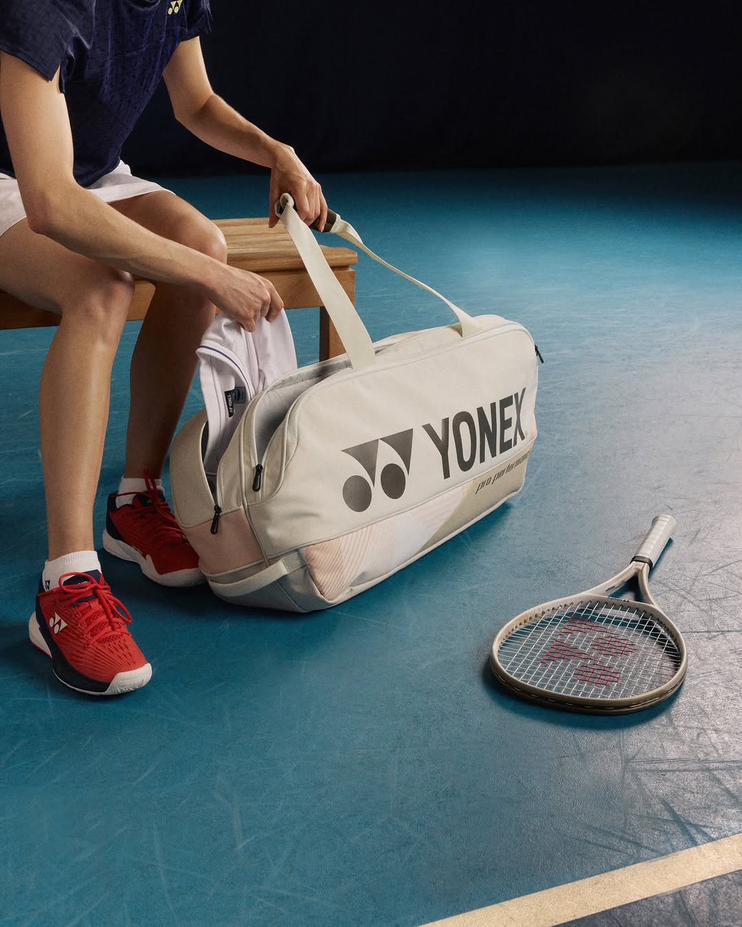 Person sitting on a bench with a Yonex bag and tennis racket on a blue court photo by Yonex