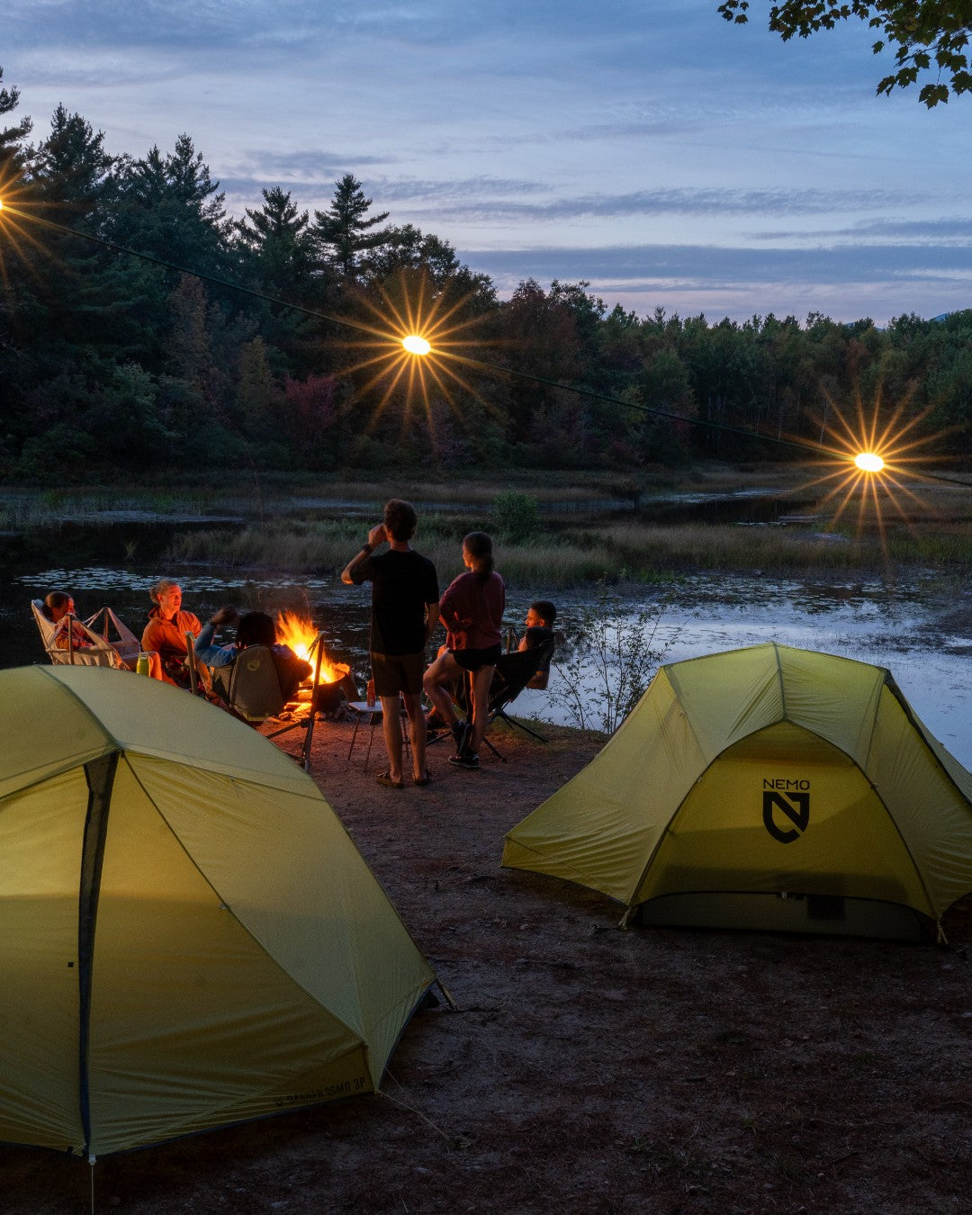 Camping scene with yellow tents, people around a fire, and a forested area at dusk. photo by nemo