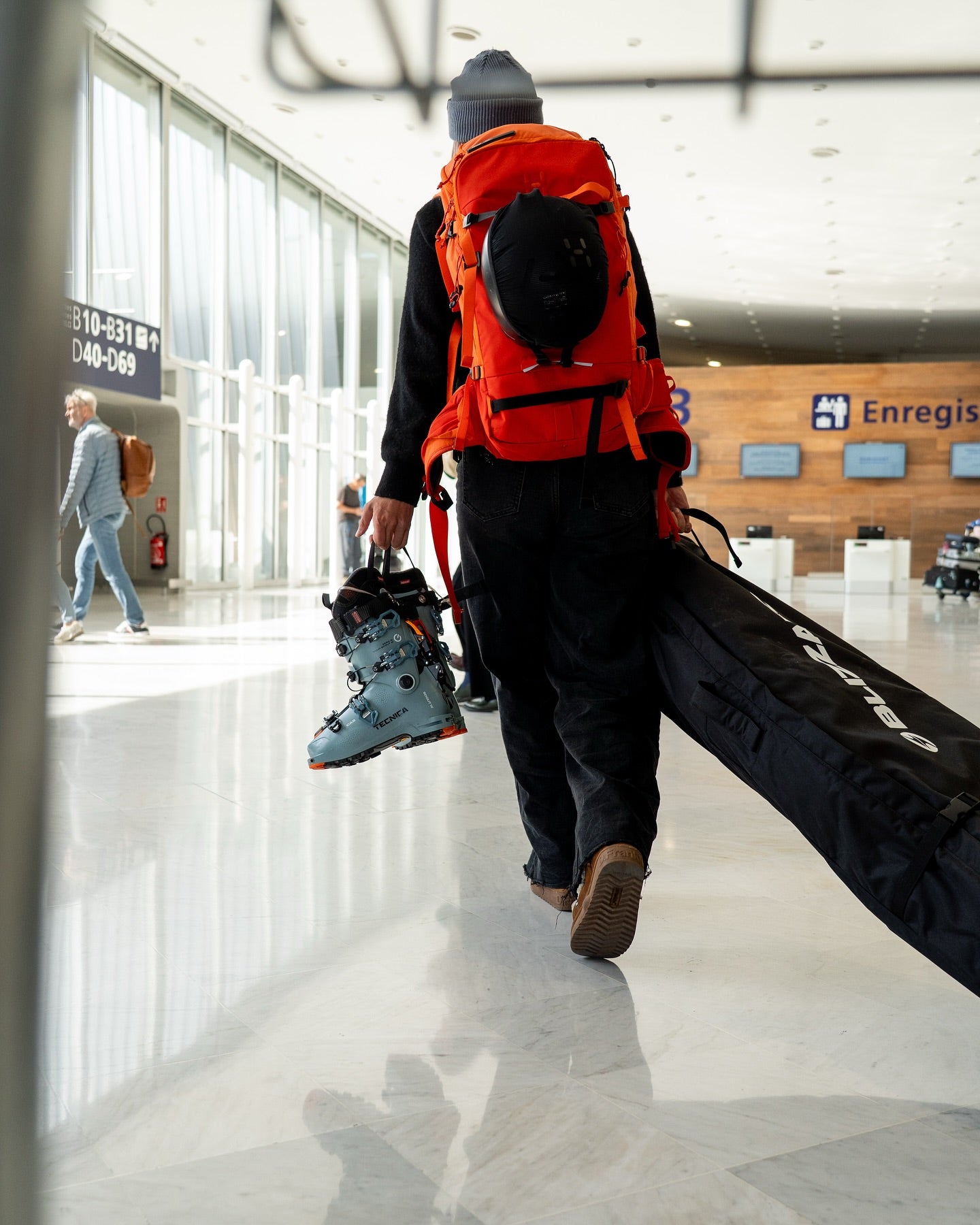 Person with an orange backpack and ski equipment walking through a modern building. photo by tecnica