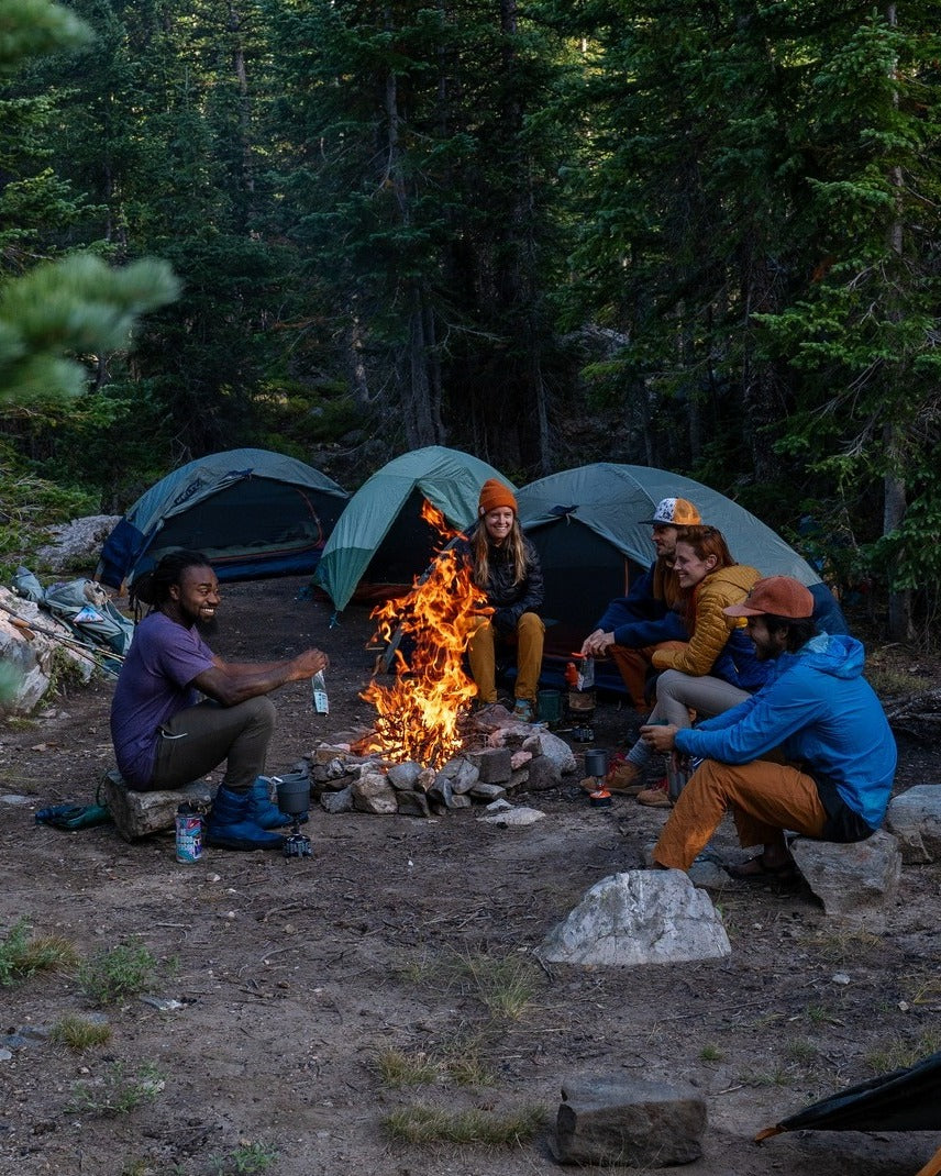 A group of four campers sits around a fire pit at a forest camping site with three green tents in the background.