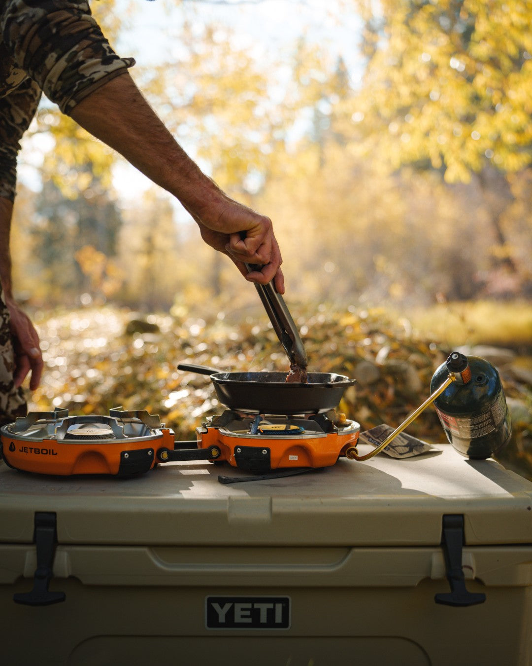 Person cooking outdoors using a portable stove on a YETI cooler with autumn scenery. photo by Jetboil