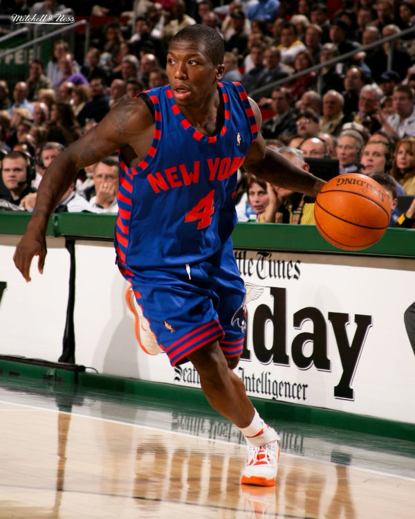 Basketball player in a blue and red New York jersey dribbling a ball on a court with spectators in the background.