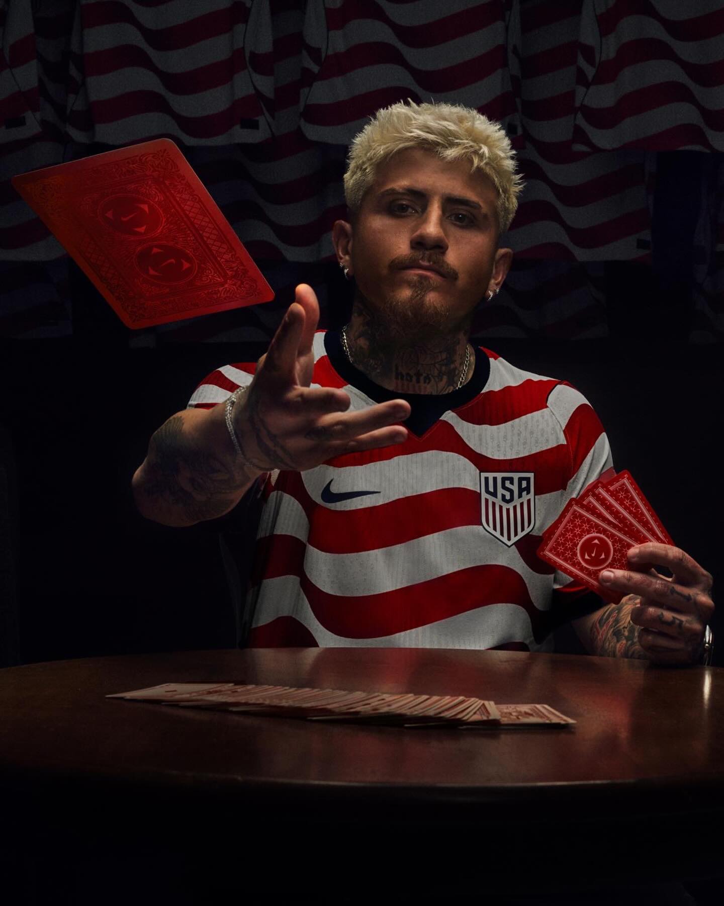Pro soccer player in a red and white sjersey with a USA logo holding playing cards against a dark background.
