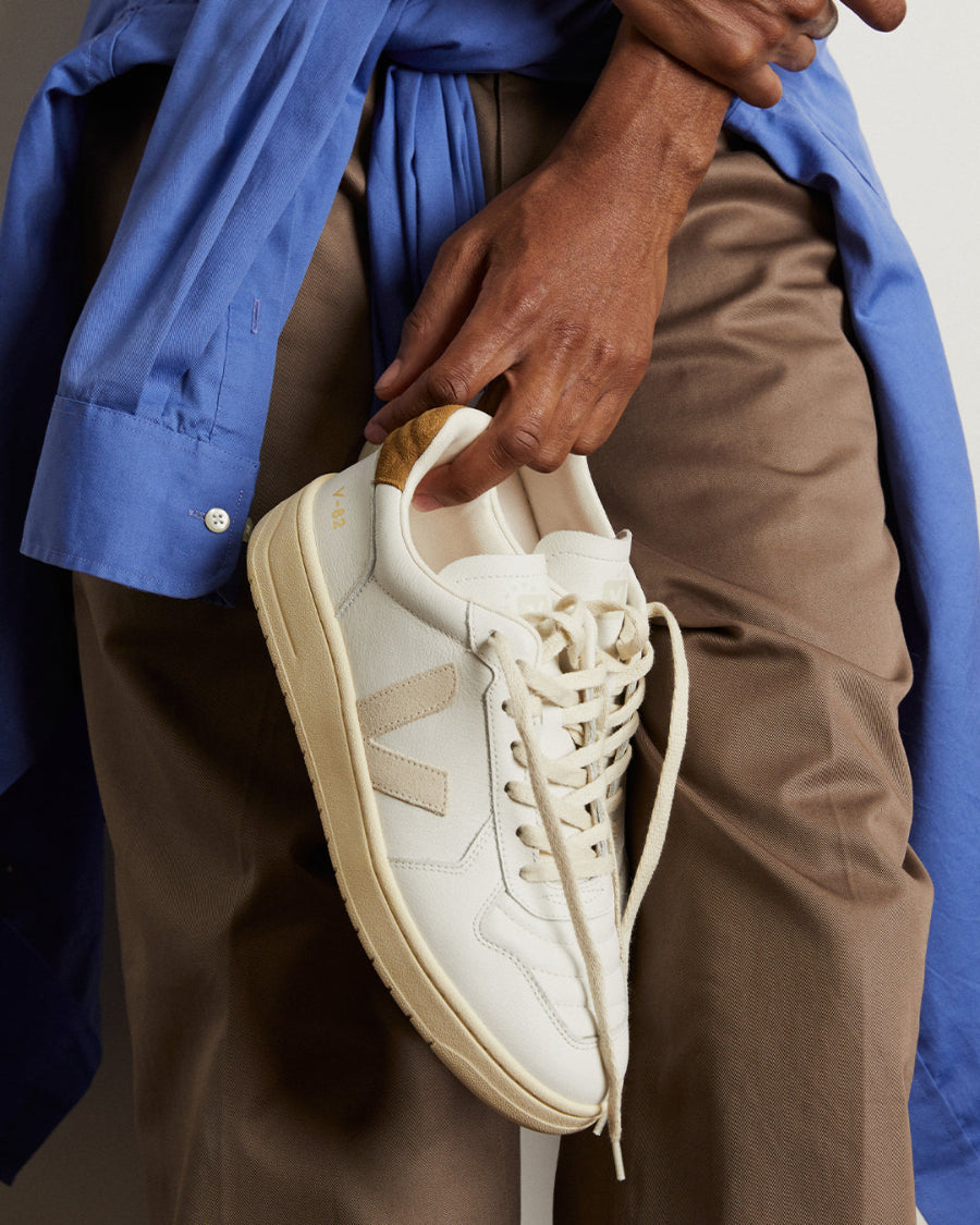 A close-up of a person holding a white veja leather sneaker with beige side stripes and a light wood heel pull tab, styled with a blue shirt and brown pants.