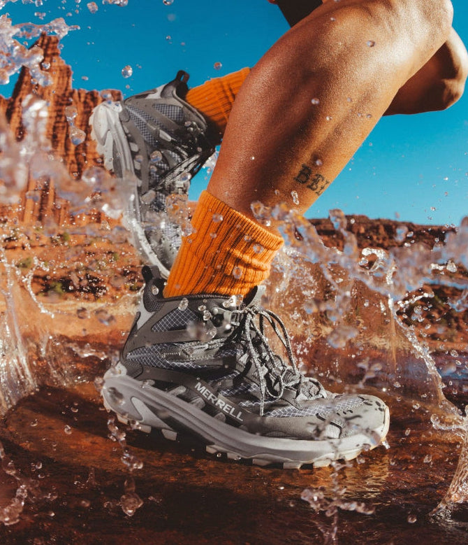 A close-up action shot of a person's leg running through splashing water outdoors, wearing a gray merrell hiking boot, bright orange crew socks, and black shorts against a blue sky and red rock desert background.