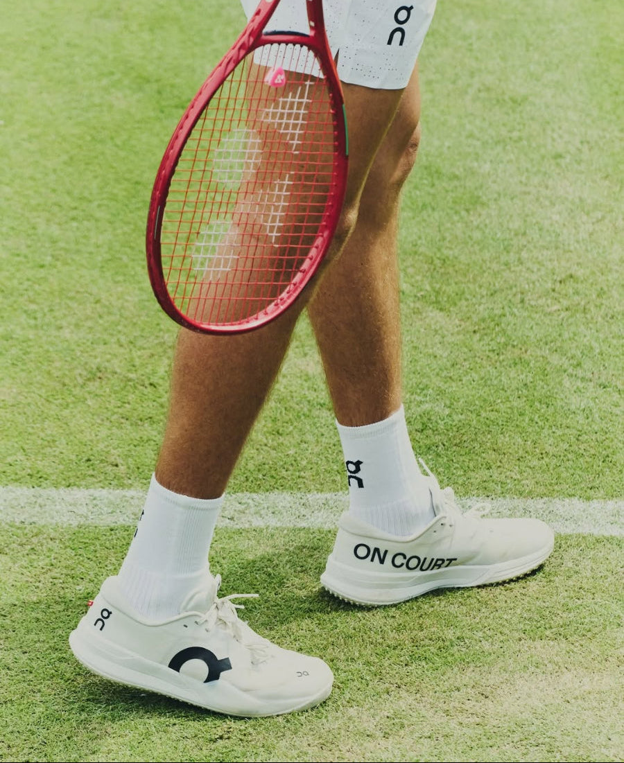 A close-up of a tennis player's legs on a grass court, wearing white On Court athletic shoes with white socks and holding a red tennis racket.