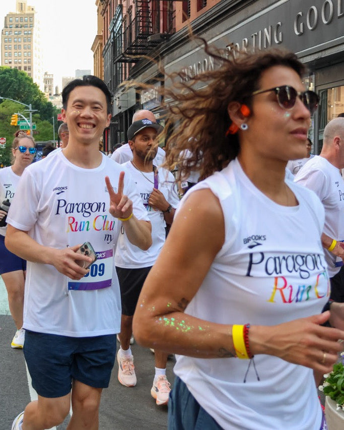 A group of runners participating in a Paragon Run Club's annual pride run with a woman in the foreground wearing a white 'Paragon Run Club' tank top and sunglasses, surrounded by other participants and urban street scenery. Photo by Kris Lowe