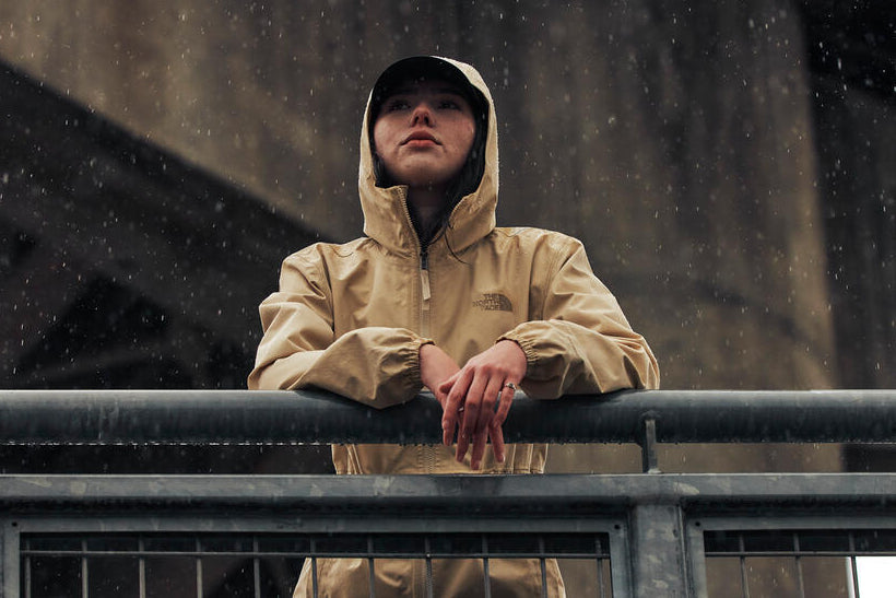 Person wearing a beige raincoat standing on a bridge during a rainy day.