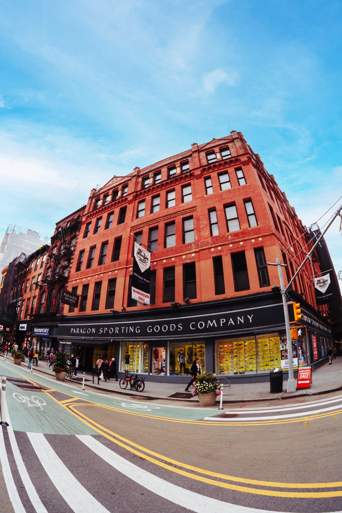 A low-angle, wide-angle street photograph of a red brick building housing the Paragon Sporting Goods  store on a the corner of 18th street and broadway