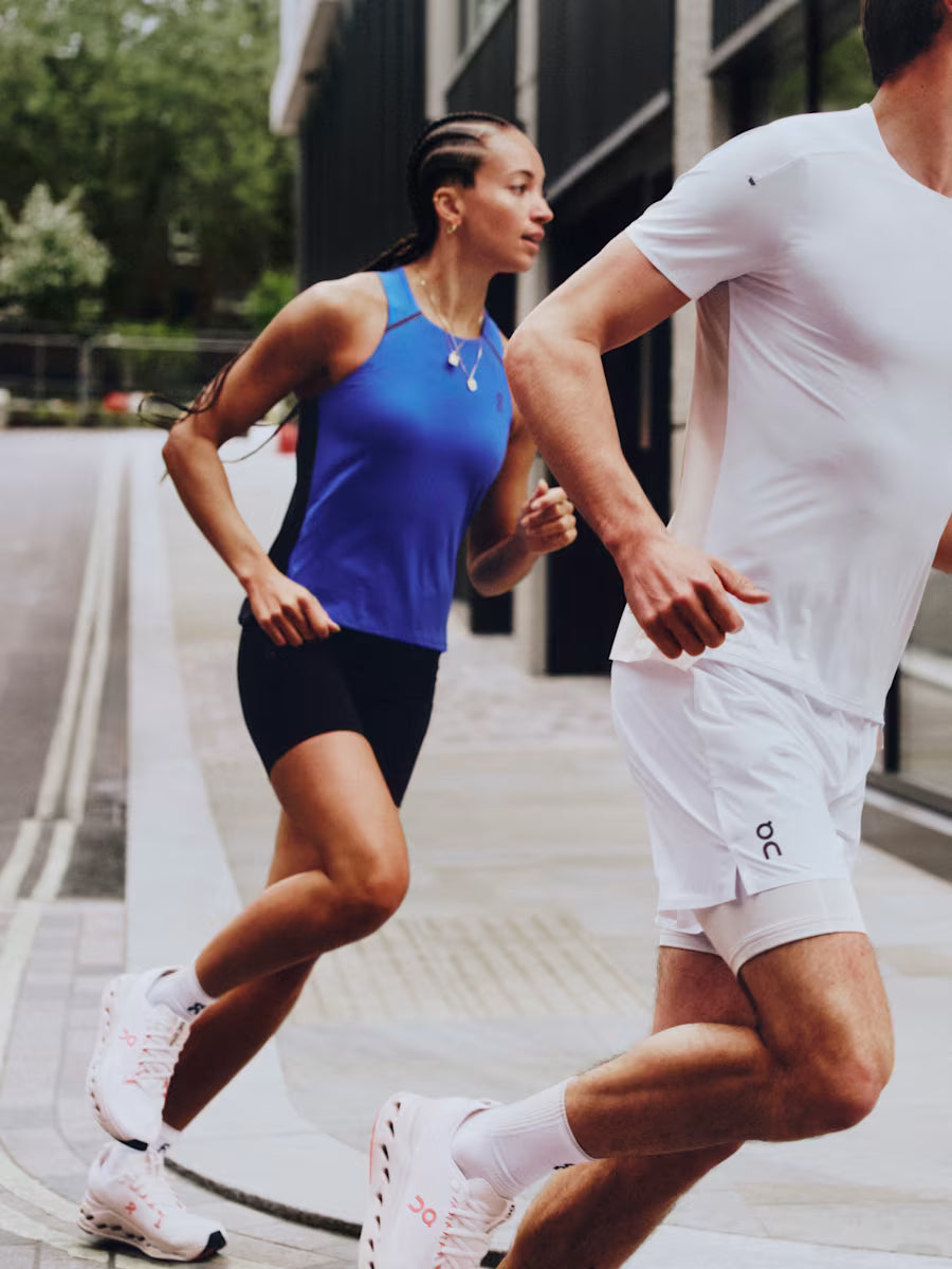 Two runners, a woman in a blue top and black shorts, and a man in a white t-shirt and shorts, running outdoors. wearing On Running Apparel photo by: On
