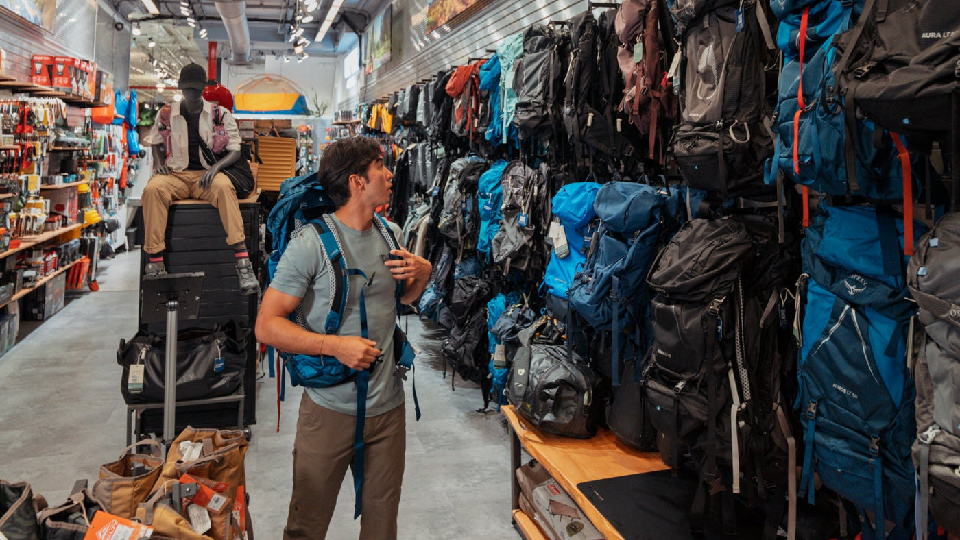 A man wearing a gray t-shirt and brown pants stands in  Paragon Sports' camping shop aisle, holding a small bottle and looking at a blue backpack hanging on the wall. The store is filled with hiking backpacks, shelves, and outdoor supplies.