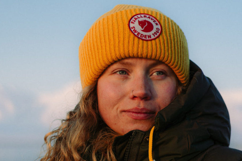 Person wearing a yellow beanie and dark jacket with a logo, standing against a blue sky. Photo by Fjallraven