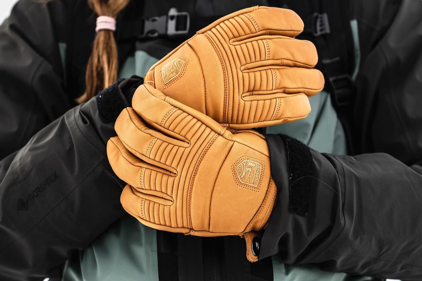 Person wearing tan gloves with a Hestra logo, holding them in front of a black jacket. Photo by Hestra