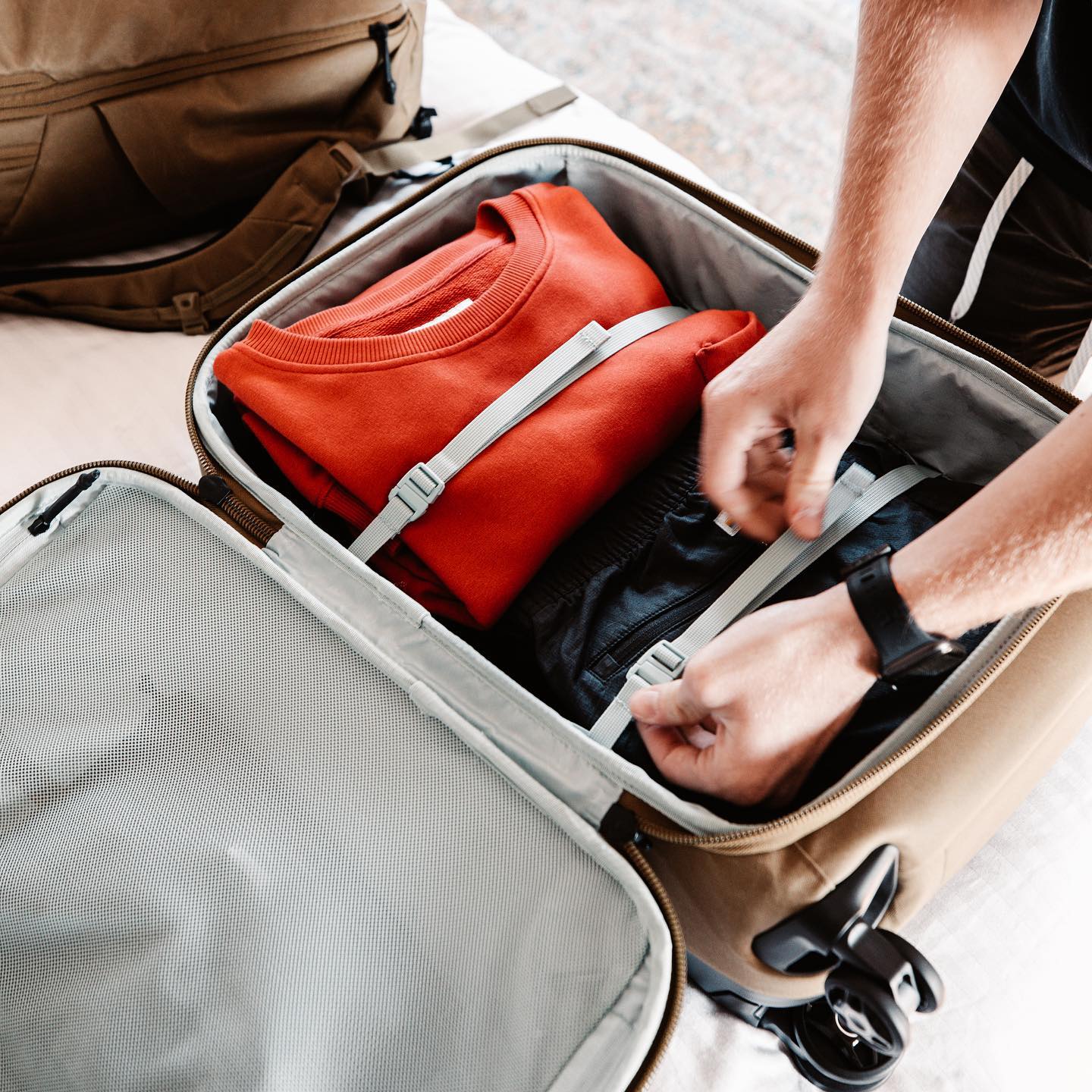 Person packing clothes into a suitcase with a backpack nearby.