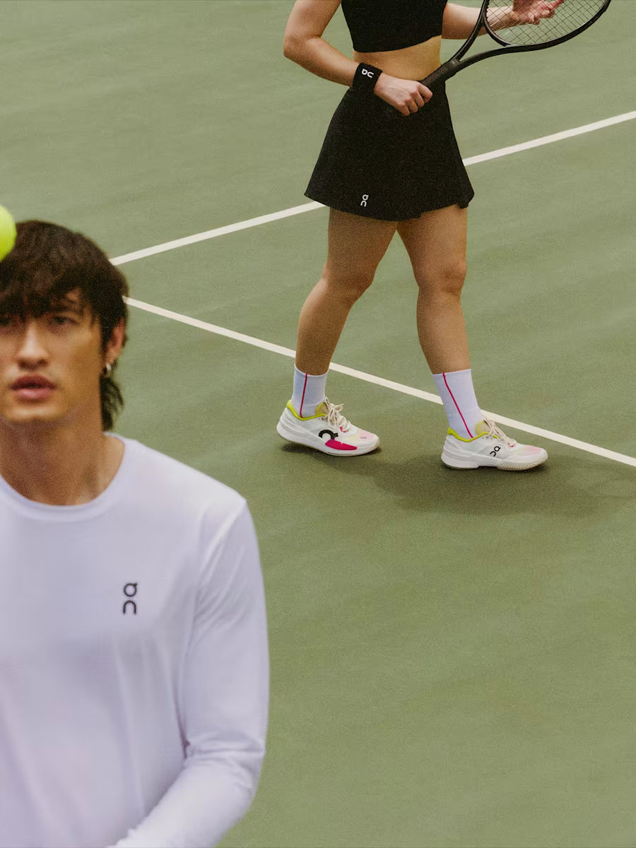 Person on a tennis court holding a racket and a tennis ball, with another person in the foreground.