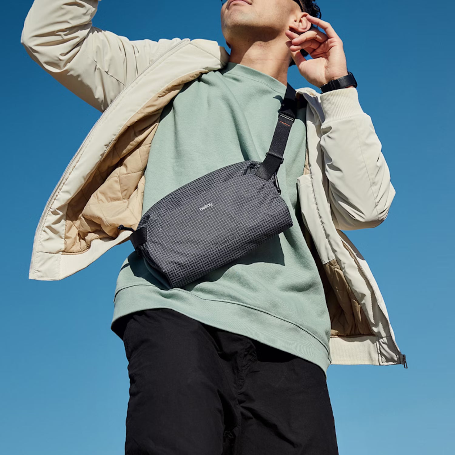 Person wearing a green shirt and beige jacket with a gray crossbody bag against a clear blue sky. photo by bellroy
